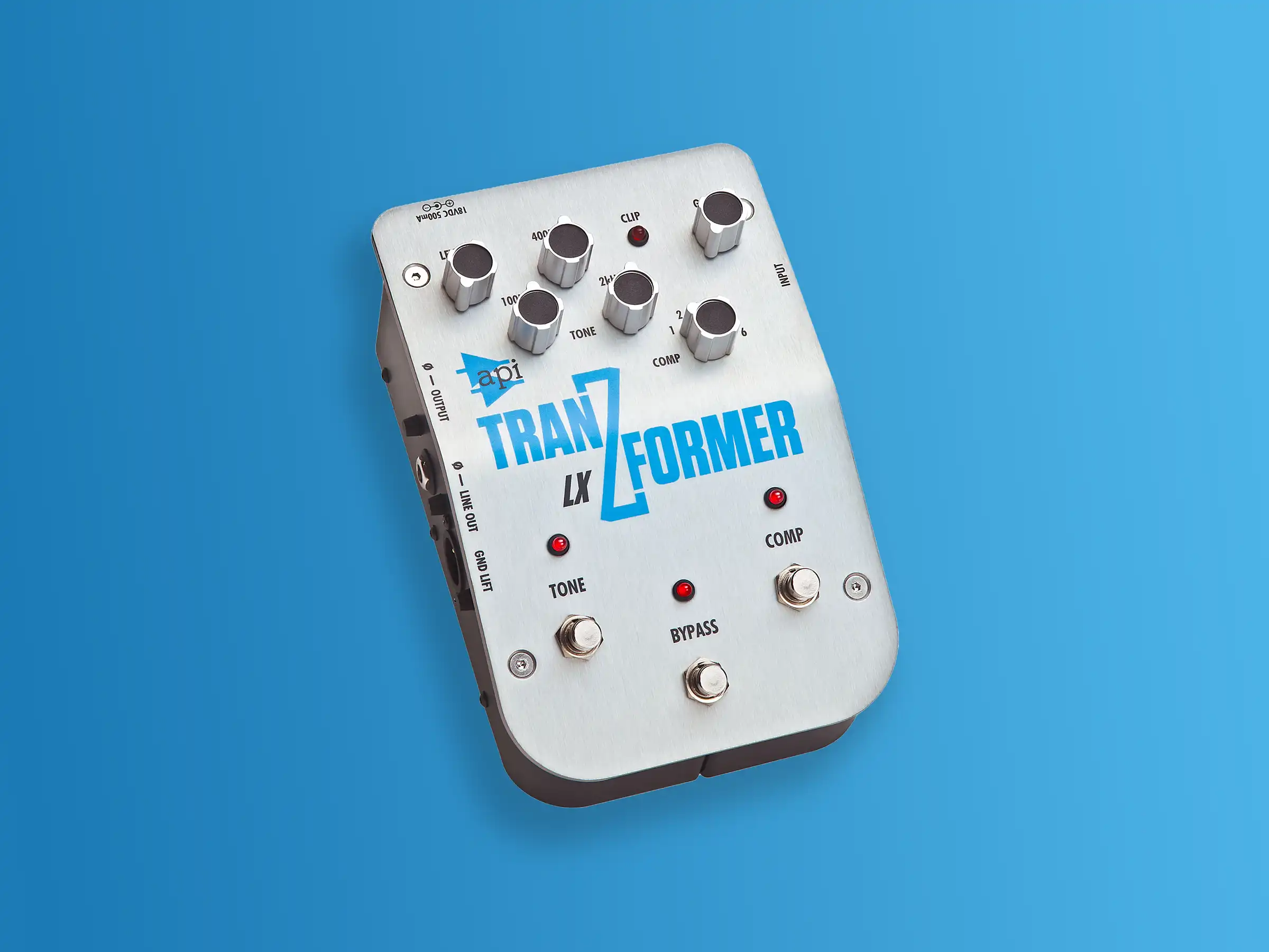The API Audio TranZformer LX bass pedal, a silver stompbox with blue lettering, rests on a solid blue background. It features multiple knobs for tone and compression control, along with input and output jacks, and a bypass footswitch. The pedal's design is sleek and modern.
