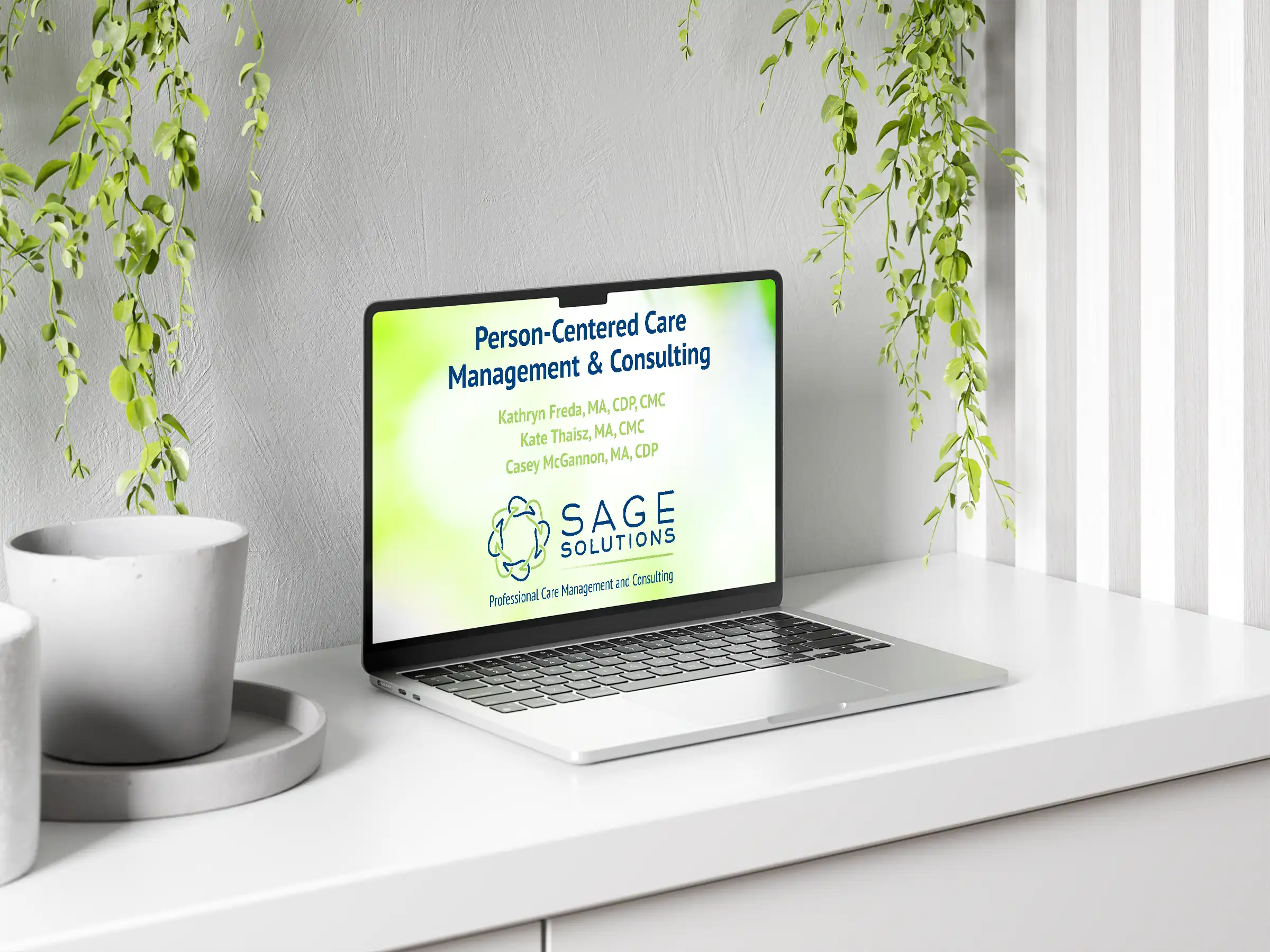 A laptop displaying the Sage Solutions website on a white desk with a gray wall and hanging plants. The website title is "Person-Centered Care Management & Consulting" and lists team members.